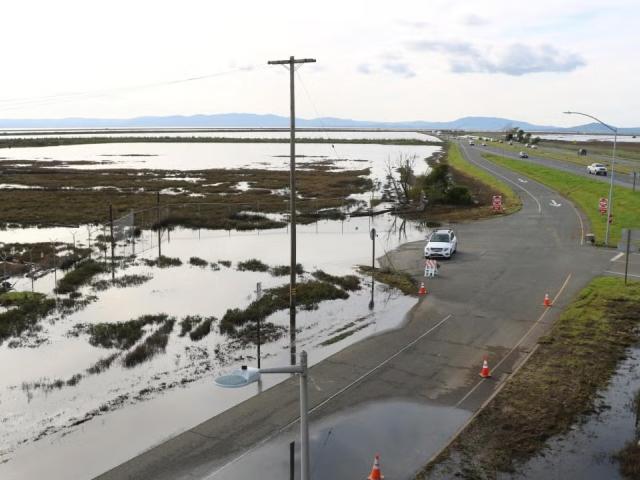 Coastal road beside flooded marsh, parked car and utility pole, distant hills under clouds