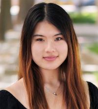 Headshot of a young woman with long dark-to-brown hair, soft smile, blurred outdoor background